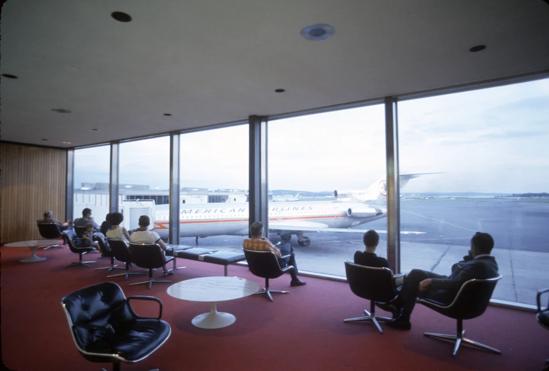 People sit in a 1960s airport lounge with red carpet, looking out large windows at an American Airlines jet parked at the gate under a cloudy sky.