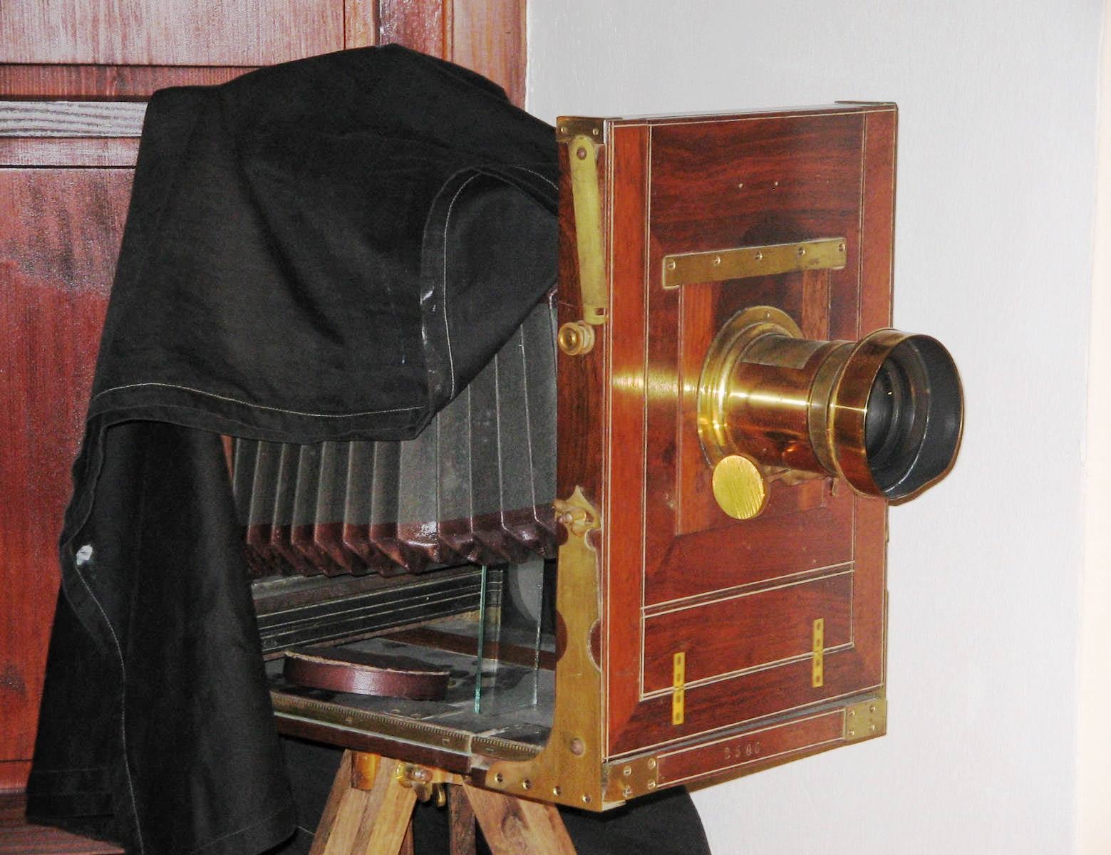 A vintage large format wooden camera with brass fittings and a black cloth draped over the back, positioned on a tripod.