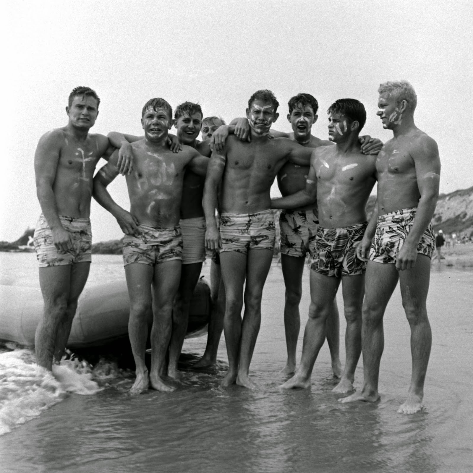 Seven young men in patterned swim trunks stand together on a beach, smiling and posing with arms around each other. Some have sunscreen on their faces. An inflatable raft and waves are visible at their feet.