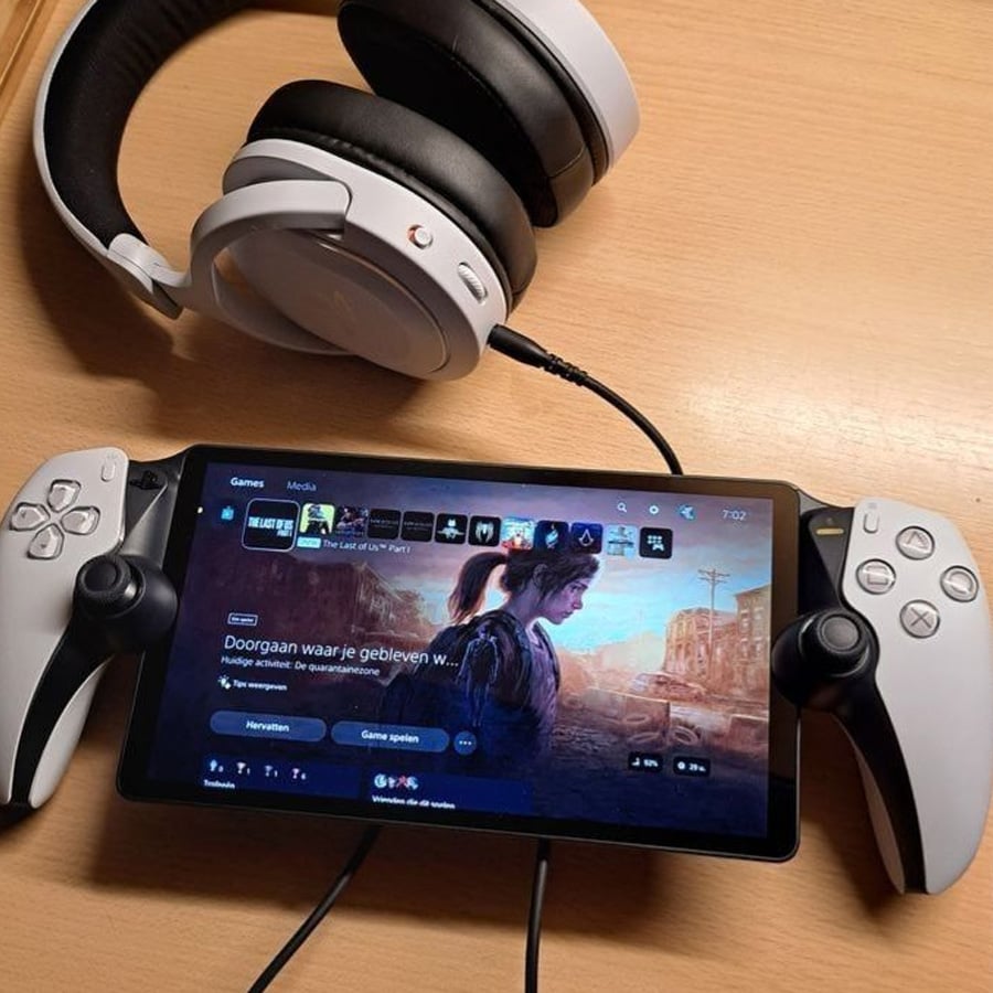 A tablet with two gaming controllers attached displays a game selection screen. A pair of over-ear headphones rests nearby on a wooden desk.