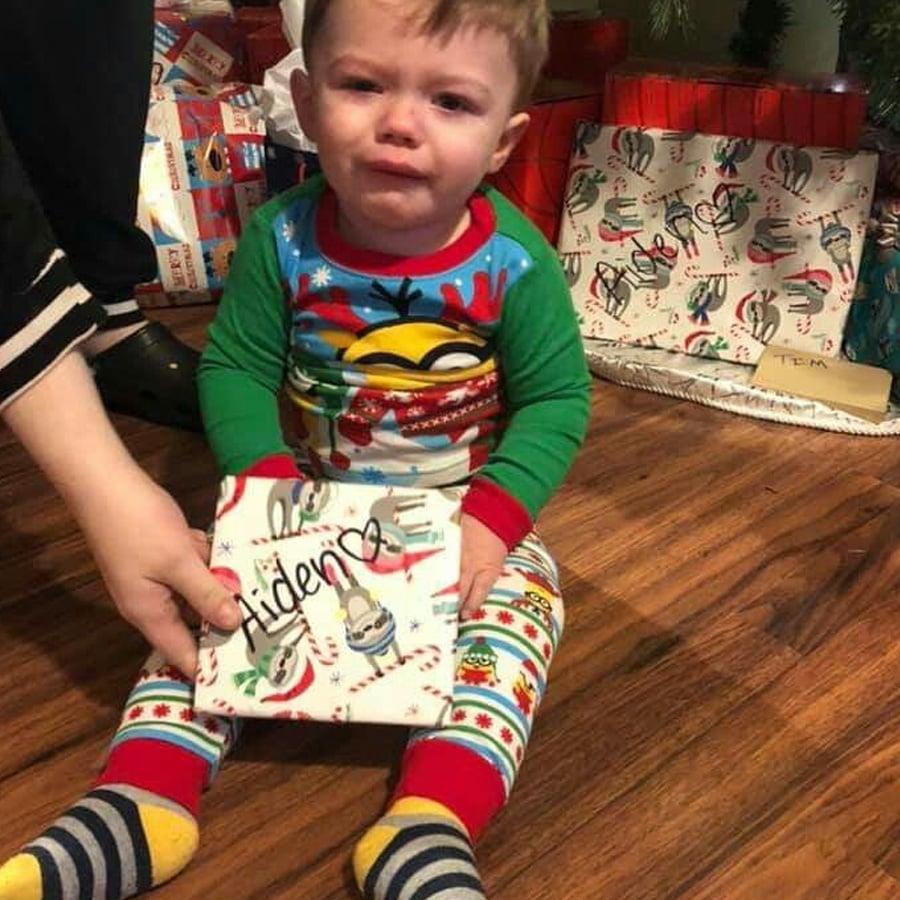 A young child in colorful pajamas sits on the floor, crying, while holding a wrapped gift labeled “Aiden.” Presents and a decorated tree are in the background. An adult hand is gently touching the gift.