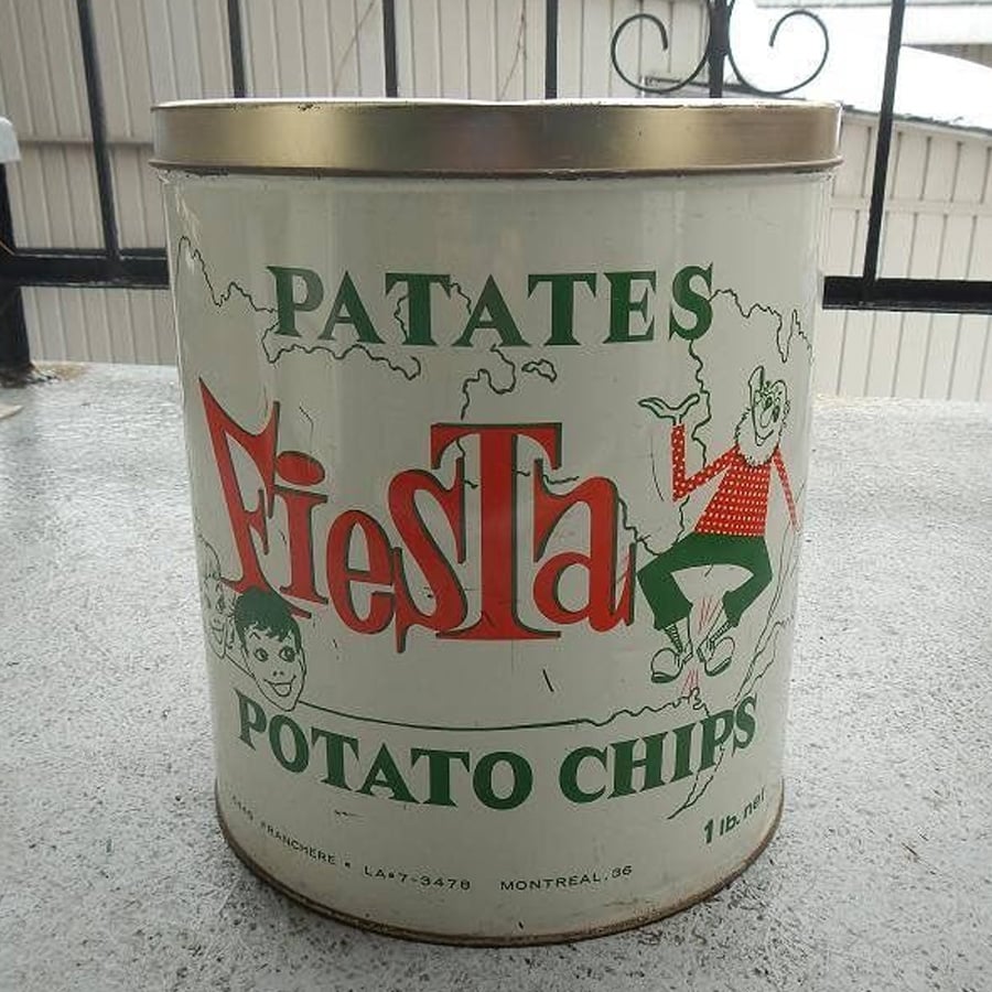 A vintage tin can with “Patates Fiesta Potato Chips” written on it, featuring cartoon figures and a festive, retro design. The can is set on a concrete surface, with a building and railing in the background.