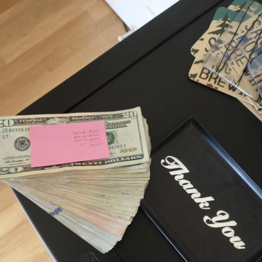 A stack of $20 bills held together with a pink slip sits next to a black tray labeled “Thank You” on a dark table. Part of a wooden floor and some decorative items are also visible.
