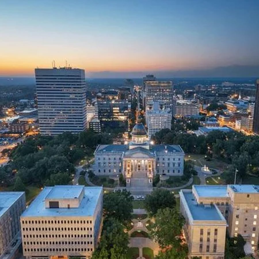 Aerial view of a city at sunset, featuring a domed government building surrounded by trees and modern office buildings, with city lights beginning to illuminate.