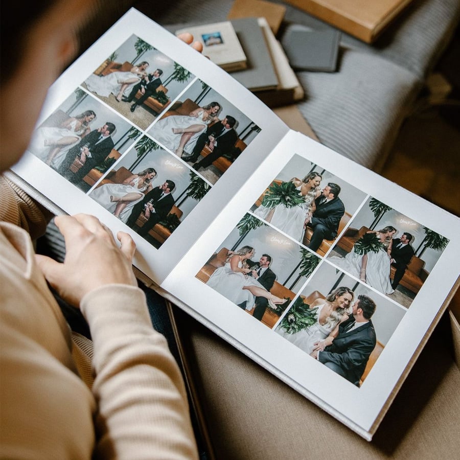 A person holds and looks at an open photo album showing several wedding photos of a bride and groom together. Other closed photo albums are stacked on a couch nearby.