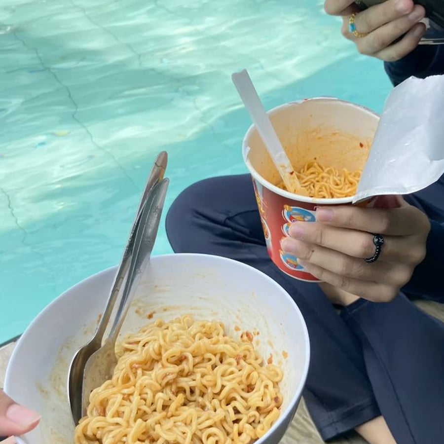 Two people sit by a pool holding instant noodle cups, one with a bowl and spoon, the other with a cup and fork. The background shows the clear blue water of the pool.
