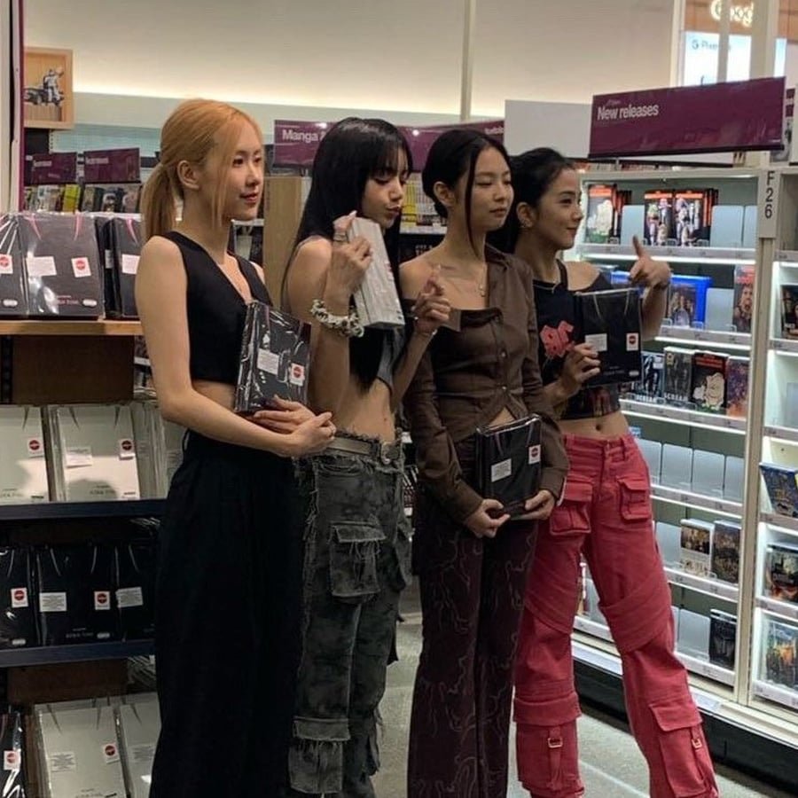 Four women stand side by side in a bookstore, holding albums and posing for a photo. They are casually dressed and standing in front of bookshelves labeled “New releases.”