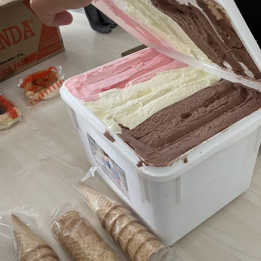 Open tub of Neapolitan ice cream showing chocolate, vanilla, and strawberry flavors. A hand lifts the lid. Ice cream cones and packaging are on the table nearby.