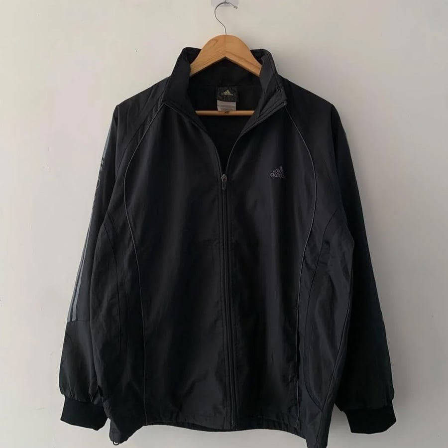 A black Adidas jacket with a zipper front hangs on a wooden hanger against a white wall. The Adidas logo is visible on the left chest area.