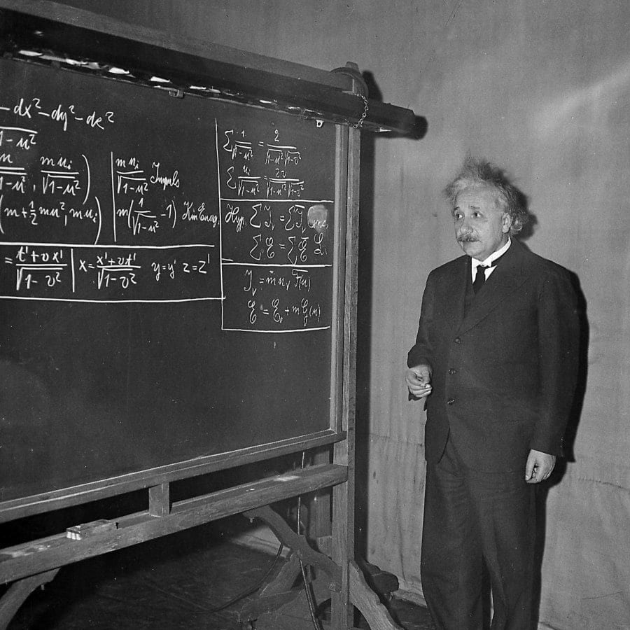 A man in a suit stands beside a large blackboard filled with complex mathematical equations, appearing to teach or explain the concepts written on the board.