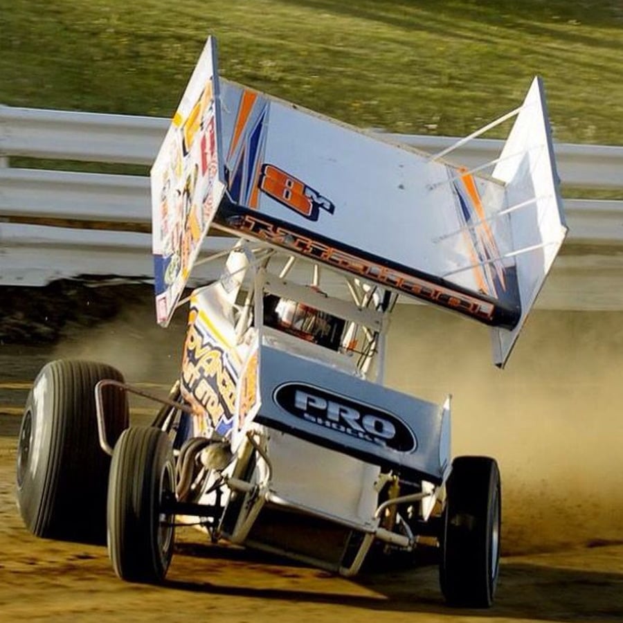 A sprint car races on a dirt track, kicking up dust as it speeds around a turn. The car has a large rear wing and colorful graphics on its body. White safety barriers line the track.