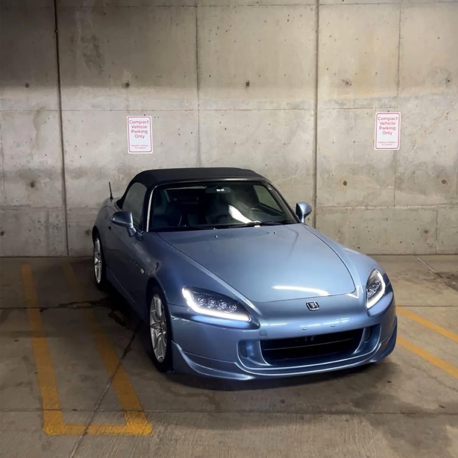 A light blue Honda S2000 sports car is parked in a concrete parking garage, partly occupying two parking spaces designated for “Compact Parking Only.” The car’s headlights and sleek design are visible.
