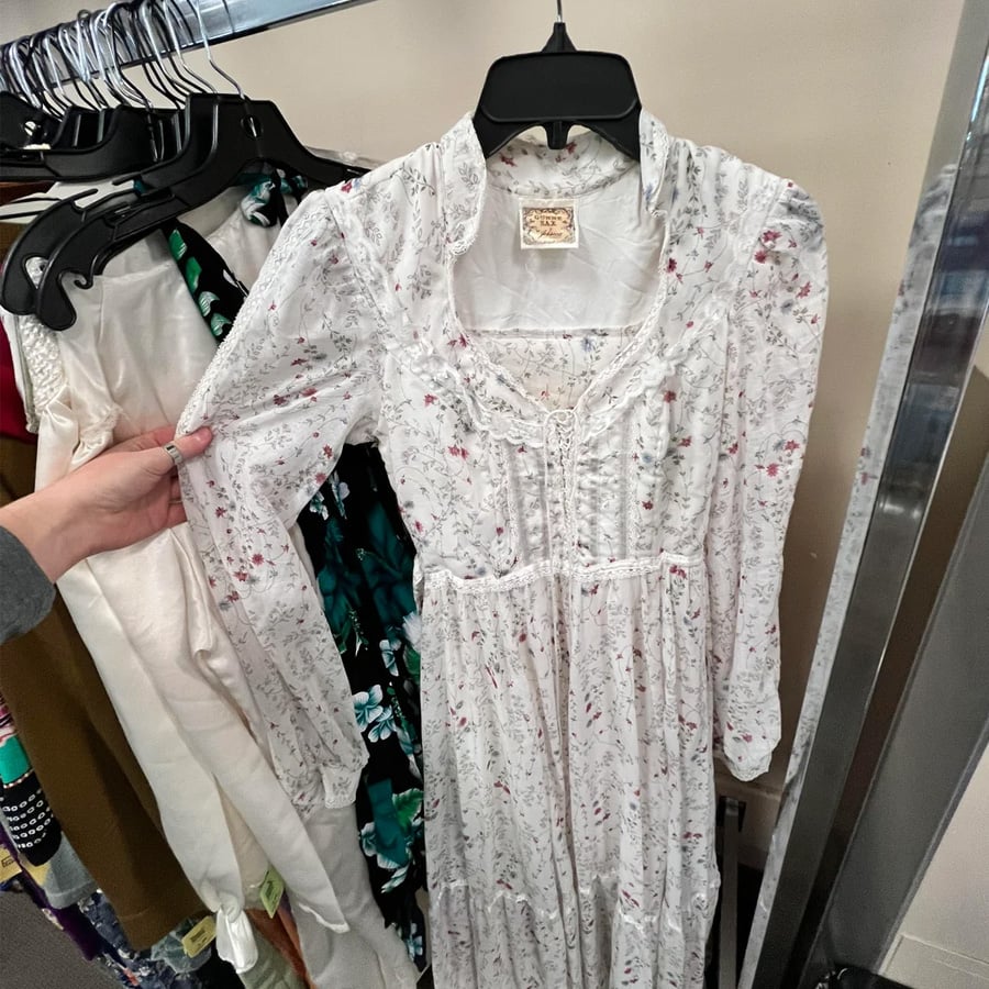 A hand holds a long-sleeve, white floral vintage dress on a hanger at a clothing rack in a store. Other assorted dresses are visible in the background.