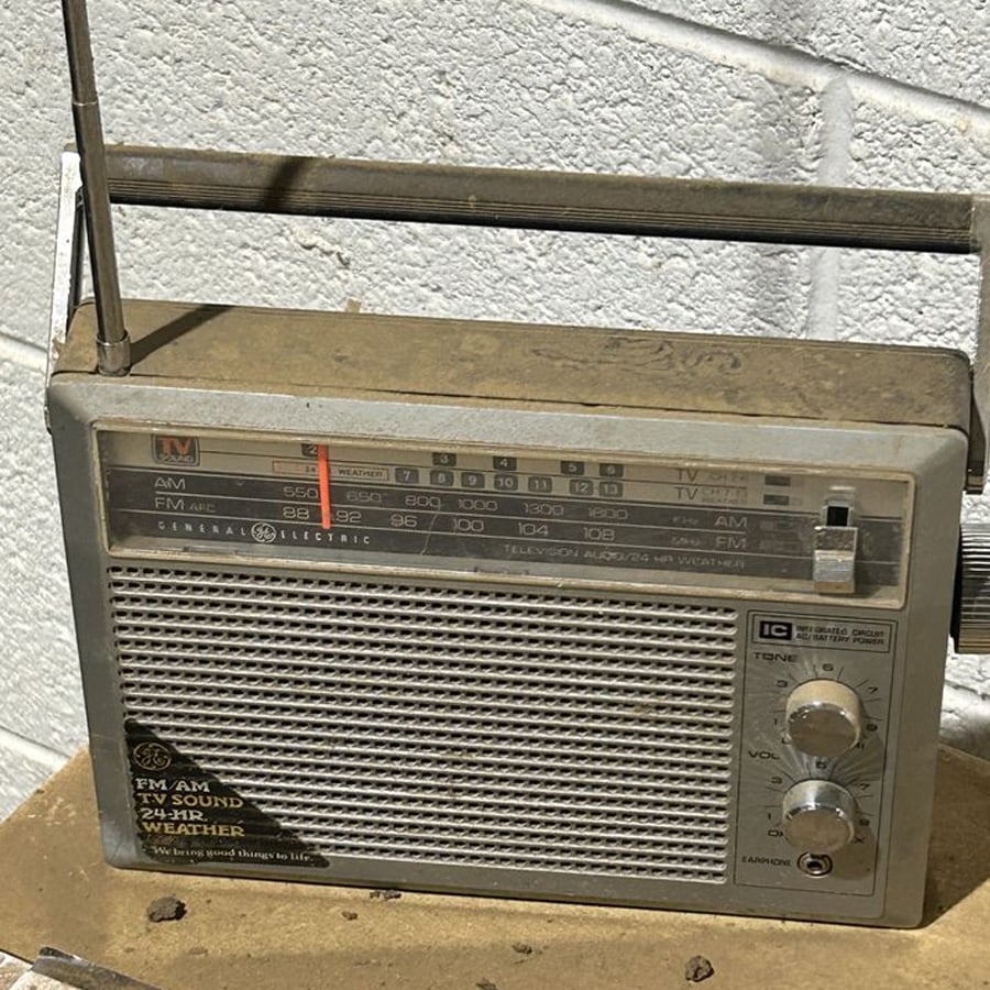 A dusty vintage portable radio with an extended antenna sits on a surface against a white brick wall. The radio has AM/FM dials, buttons, and tuning displays, and looks old and well-used.