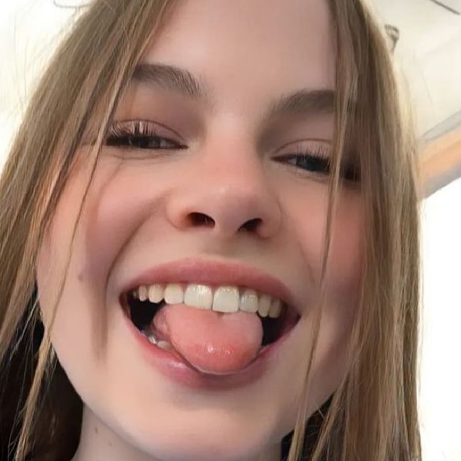 A young person with light brown hair smiles and sticks out their tongue, with hair falling over their face. The background is bright and out of focus.