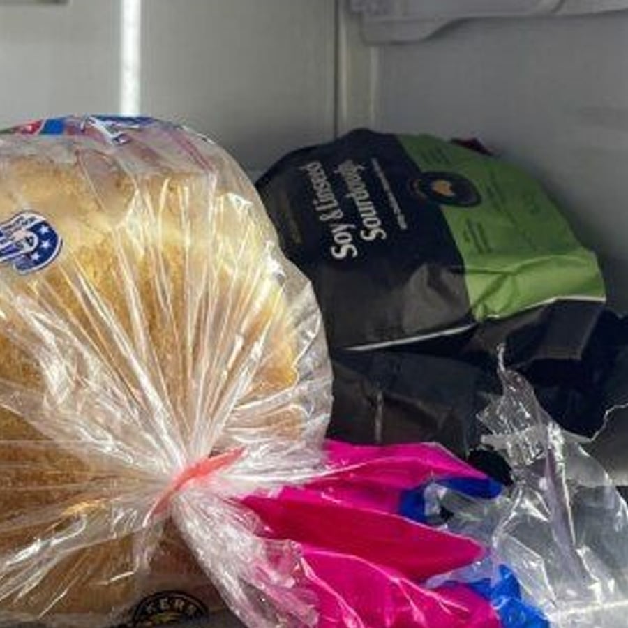 A loaf of sliced bread in a plastic bag is placed in a freezer next to a bag of soy and linseed sourdough bread. Some plastic packaging is visible in the foreground.