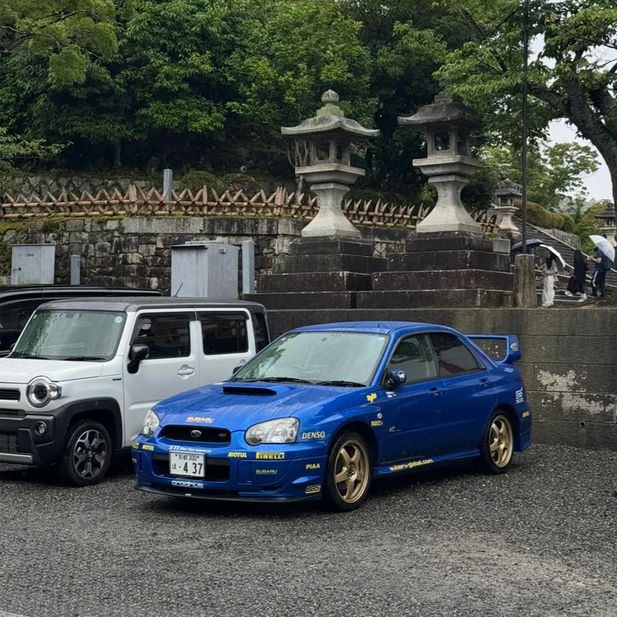 A blue sports car and a white compact car are parked side by side near stone lanterns and stairs, surrounded by greenery and a stone wall.