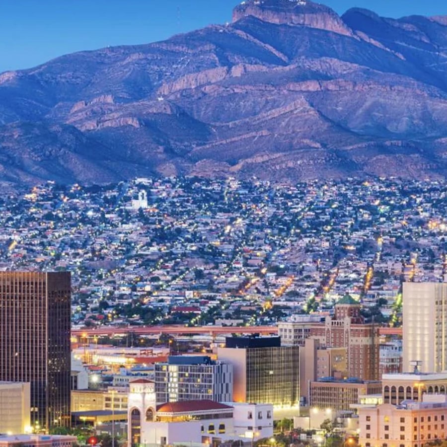 Cityscape with tall buildings in the foreground, sprawling neighborhoods, and mountains rising in the background under a clear blue sky at dusk.