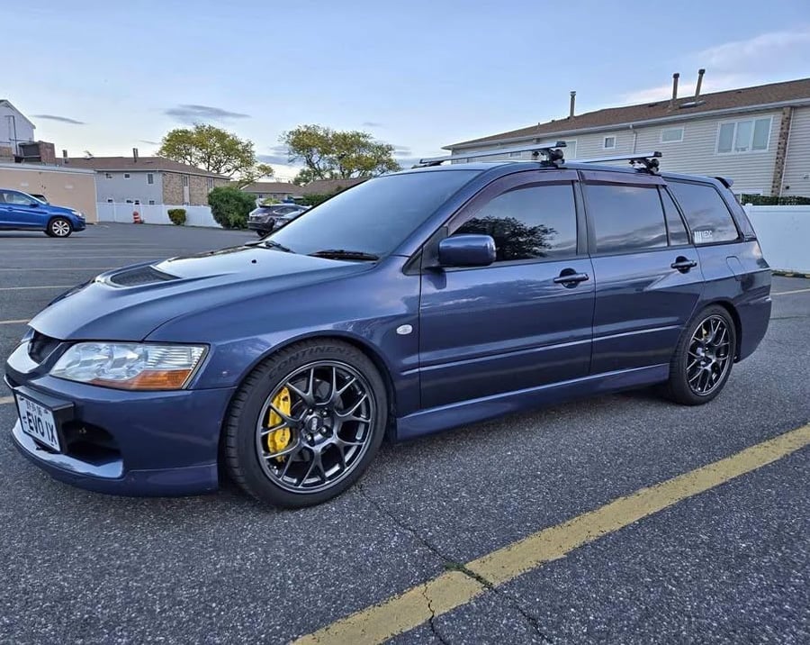 A dark blue Mitsubishi Lancer Evolution wagon with tinted windows, black wheels, and yellow brake calipers is parked in a lot near residential buildings under a clear sky.