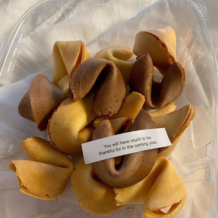 A plastic container holds several fortune cookies, some light and some dark brown. One cookie is open, revealing a fortune slip that reads, “You will have much to be thankful for in the coming year.”