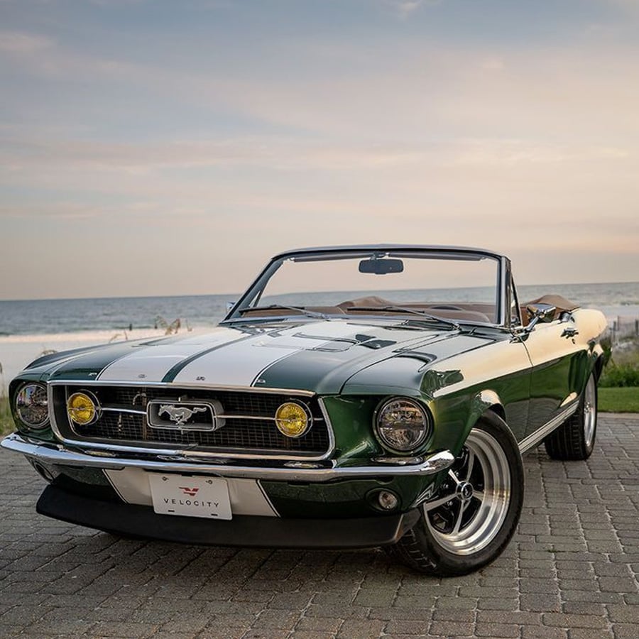 A dark green classic Ford Mustang convertible with white racing stripes is parked on a brick road by the ocean at sunset.