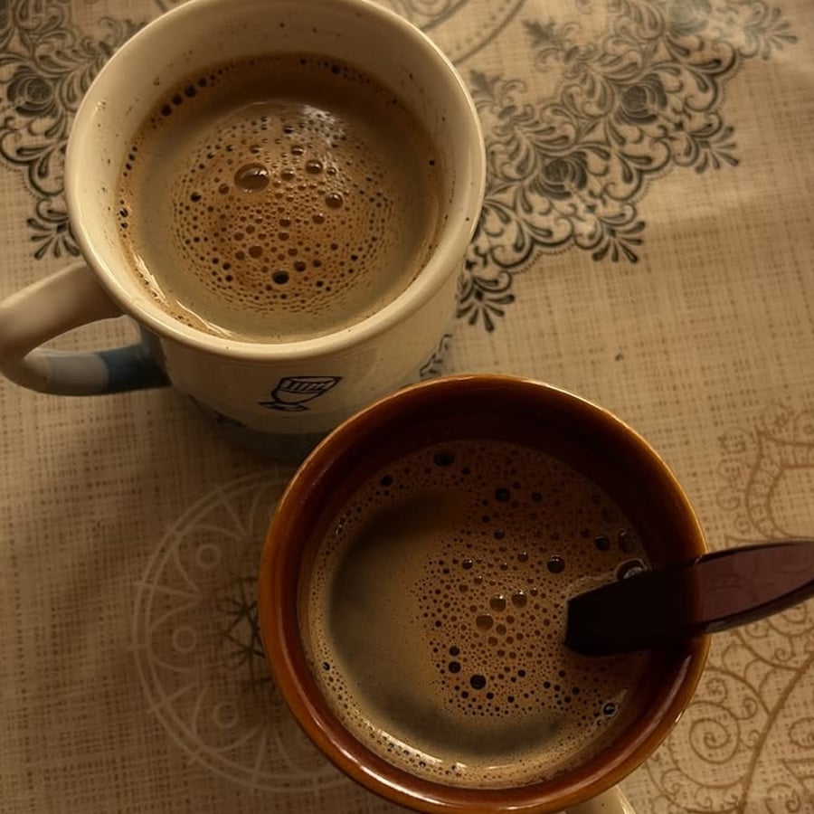 Two mugs of coffee with frothy surfaces sit on a patterned beige tablecloth; one mug has a spoon in it, and both are viewed from above.