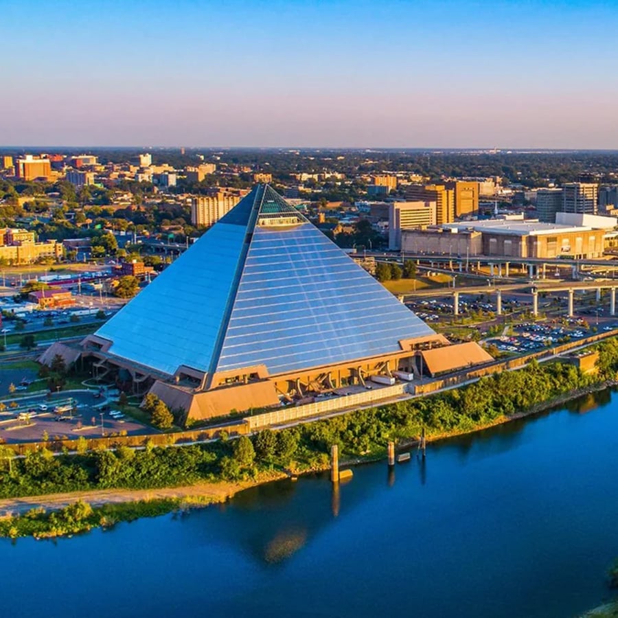 A large glass pyramid building sits beside a river, surrounded by parking lots and city buildings under a clear, blue sky.