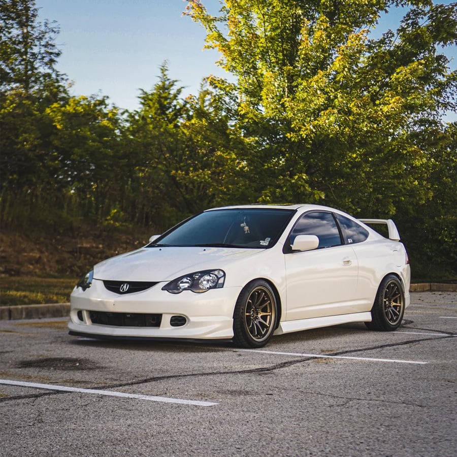 A white sports coupe with black wheels is parked in an empty outdoor lot, surrounded by green trees and shrubs under a clear sky.