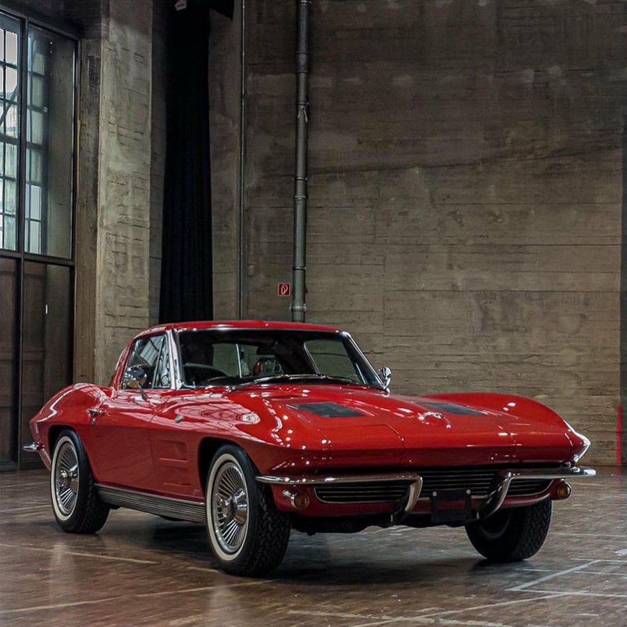 A classic red Chevrolet Corvette Sting Ray coupe is parked indoors on a wooden floor, with concrete walls and large windows in the background.