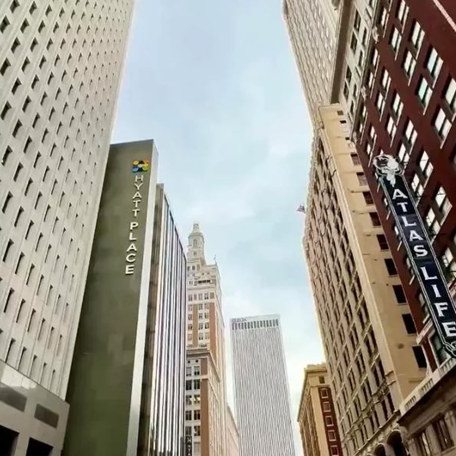Tall buildings stand close together on a city street, with signs for “Hyatt Place” and “Atlas Life.” The sky is mostly clear with some clouds.