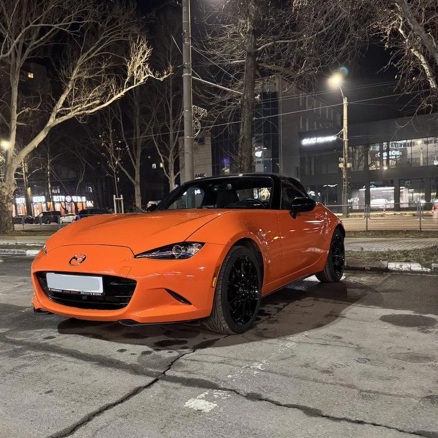 An orange Mazda MX-5 sports car is parked on a city street at night, with bare trees and illuminated buildings in the background.