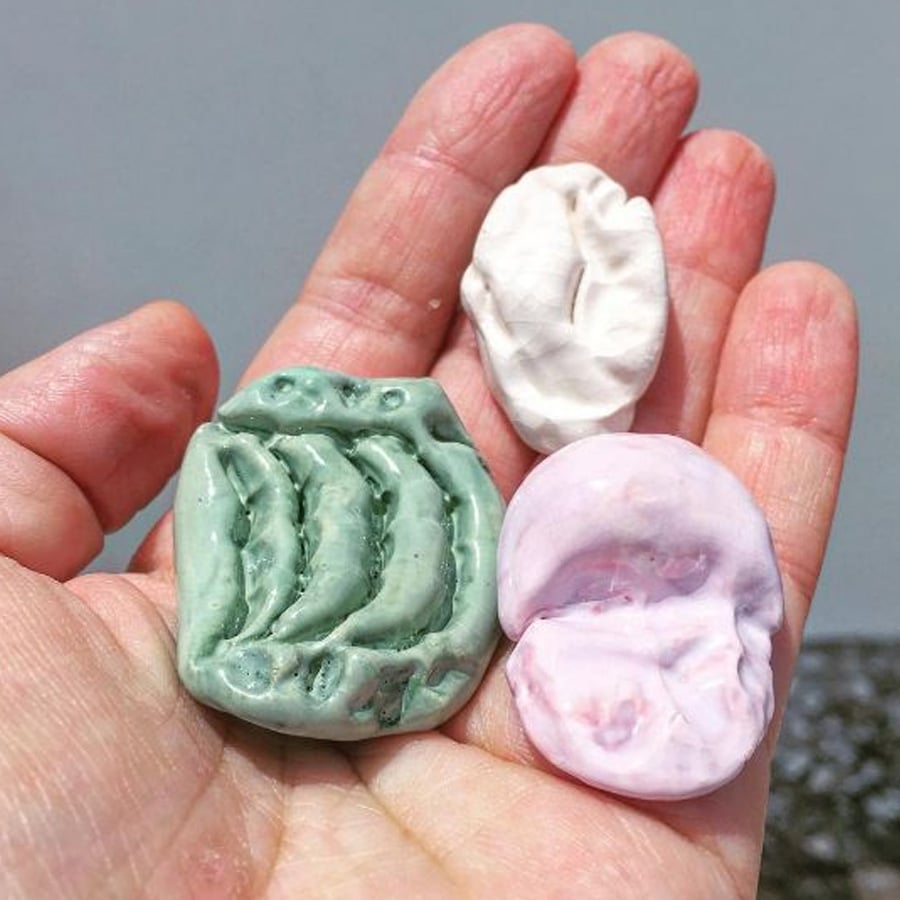 A hand holding three small, round ceramic pieces: one green with a leaf-like pattern, one plain white, and one light pink with a wavy texture.
