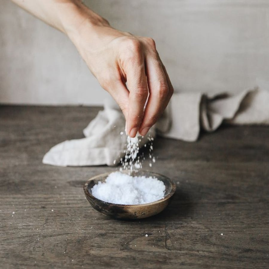 A hand sprinkles salt over a small wooden bowl filled with coarse salt, on a rustic wooden table with a beige cloth in the background.