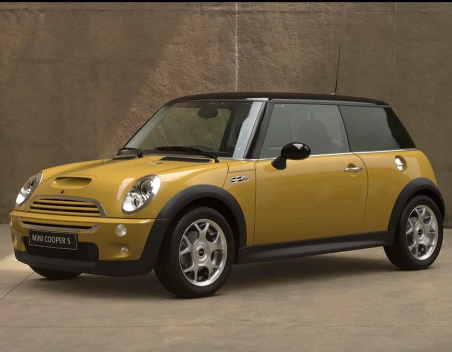 A yellow Mini Cooper S with a black roof is parked indoors against a tan background. The car is seen from the front left side, showcasing its compact design and silver wheels.