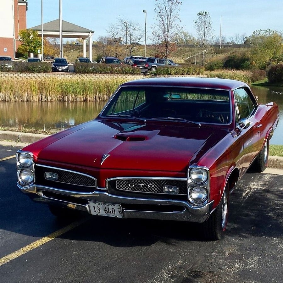 A classic red Pontiac GTO muscle car is parked diagonally on pavement near a pond, with buildings and trees in the background on a sunny day.