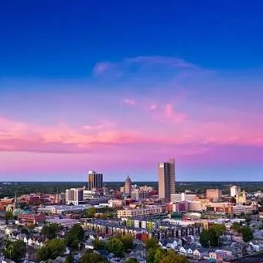 City skyline at sunset with tall buildings, colorful pink and purple clouds, and a clear blue sky above a spread of houses and greenery.