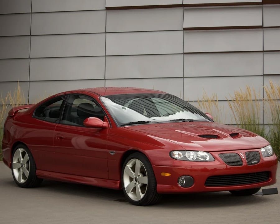 A red, two-door sports car is parked in front of a modern building with horizontal metal panels and tall grass.