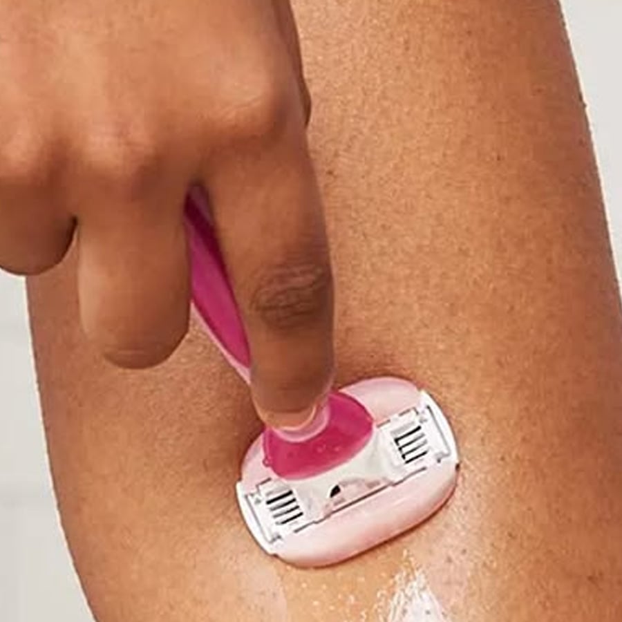 A close-up of a hand holding a pink razor, shaving a wet, soapy area on a person’s leg.