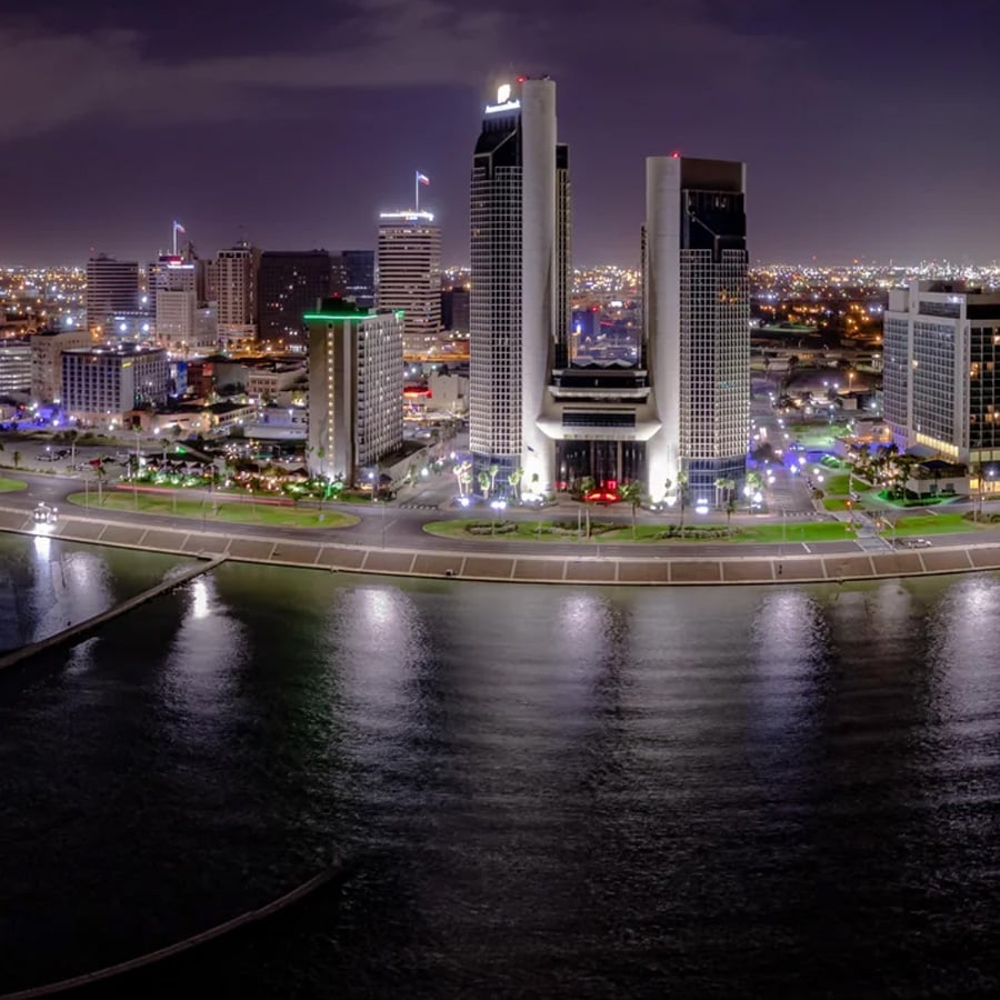 Nighttime cityscape of downtown Corpus Christi, Texas, featuring illuminated skyscrapers, waterfront buildings, and reflections of city lights on the calm bay water.