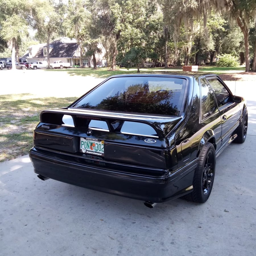 A black Ford Mustang coupe with tinted windows is parked on a driveway, surrounded by grass and trees. The car features a unique rear spoiler and a Florida license plate reading “PONY 302.”