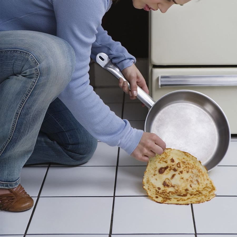 A person in a blue sweater and jeans picks up a dropped pancake from a white tiled kitchen floor while holding a metal pan.