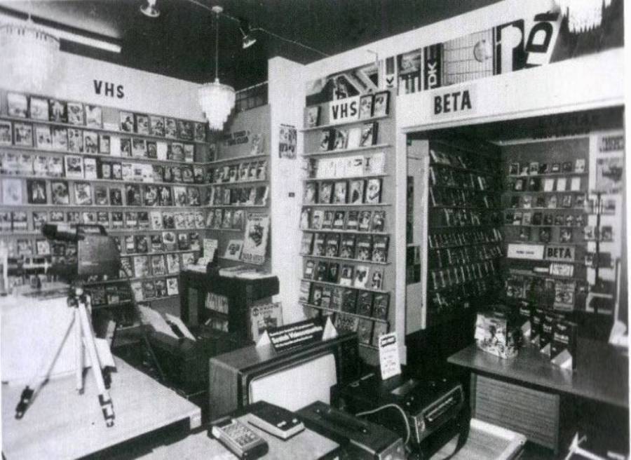 Black and white photo of a vintage video rental store with shelves labeled “VHS” and “BETA,” displaying tapes. Electronics and video players are visible on tables in the foreground.