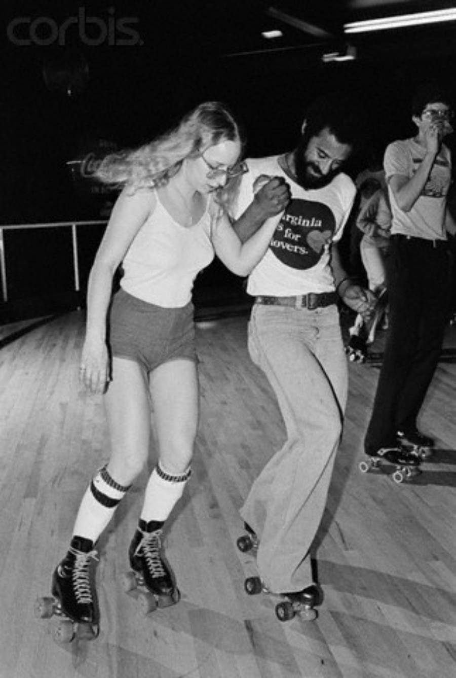 A woman and a man roller skate together, holding hands, in a skating rink. The woman wears shorts and knee-high socks; the man wears jeans and a “Virginia is for Lovers” shirt. Other skaters are visible in the background.
