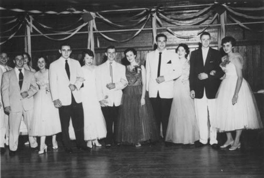 A group of young men and women in formal attire pose together at a decorated venue, likely at a prom or dance, with streamers hanging from the ceiling.