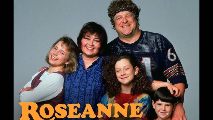 Five people pose together, smiling, with the word “Roseanne” in bold orange letters at the bottom. One man wears a sports jersey; others wear casual clothes. They appear to be a family.