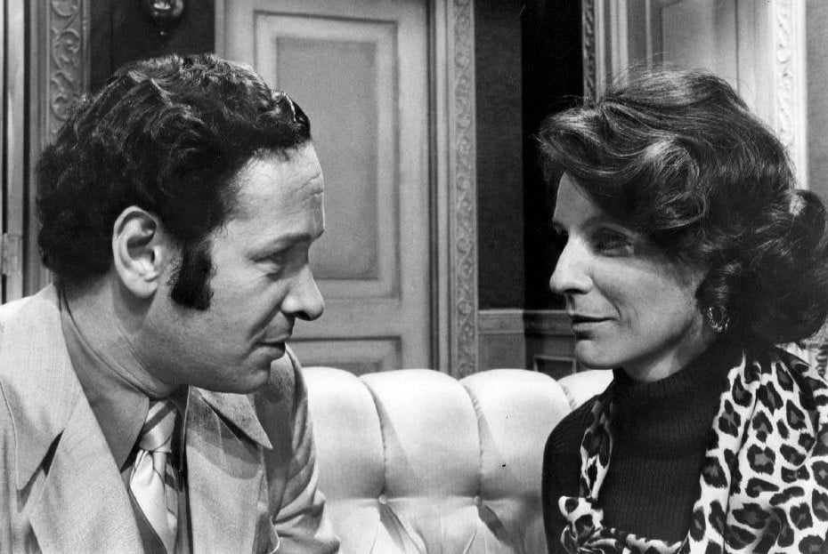 A man in a suit sits on a couch, gazing at a woman in a patterned scarf. They appear to be having a serious conversation in an elegantly decorated room. The image is black and white.