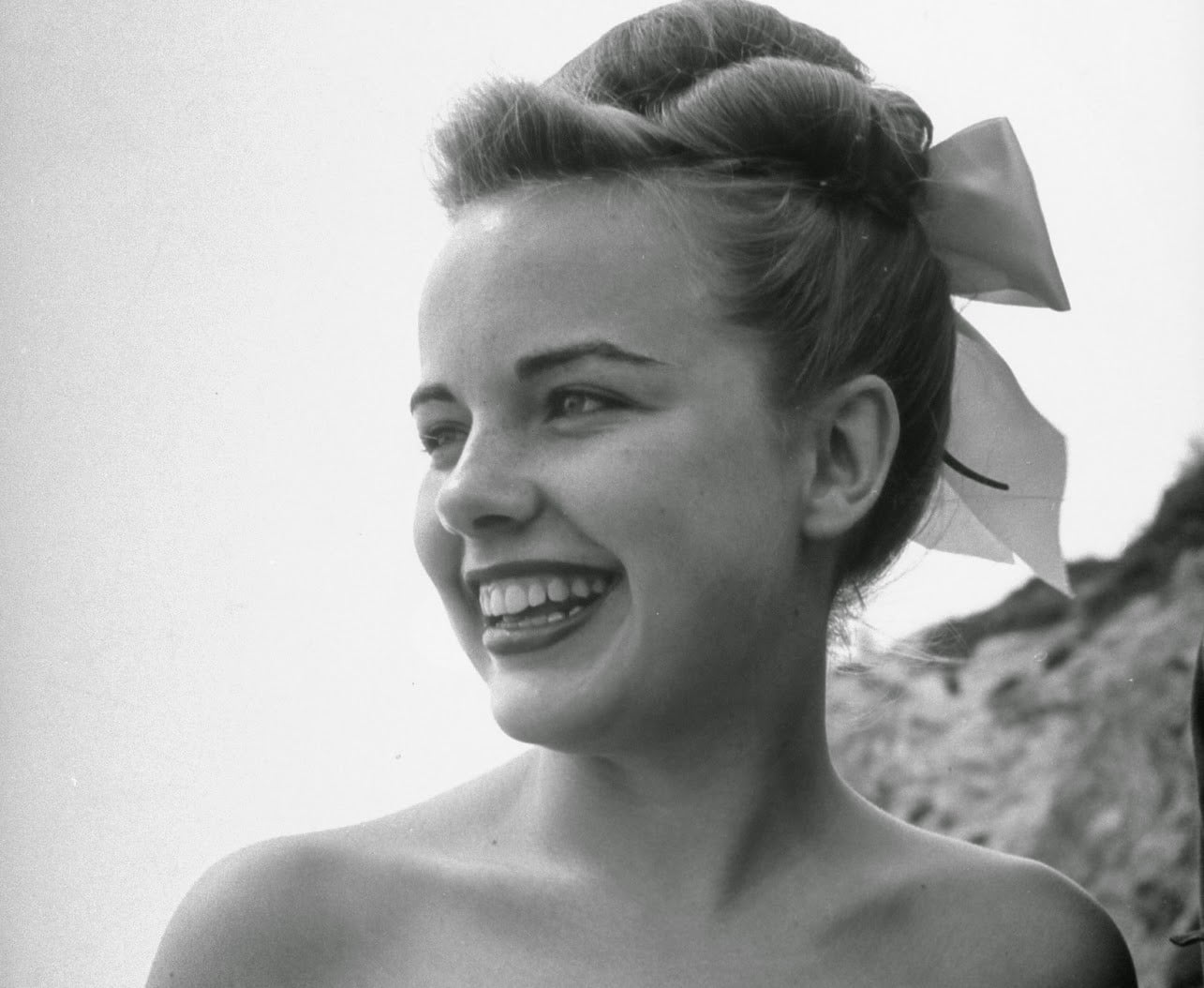 A young woman with a large bow in her hair smiles while looking to the side, outdoors, with a blurred hillside in the background. The photo is in black and white.