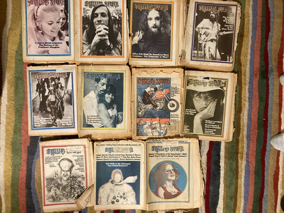 Ten vintage Rolling Stone magazine issues with black and white covers featuring various celebrities are arranged in two rows on a striped rug. The magazines show signs of age and wear.