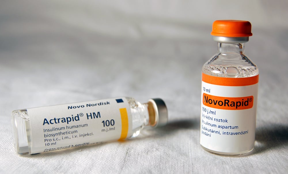 Two vials of insulin: one labeled “Actrapid HM” lying on its side and another labeled “NovoRapid” standing upright, both with white labels and orange caps, on a white surface.