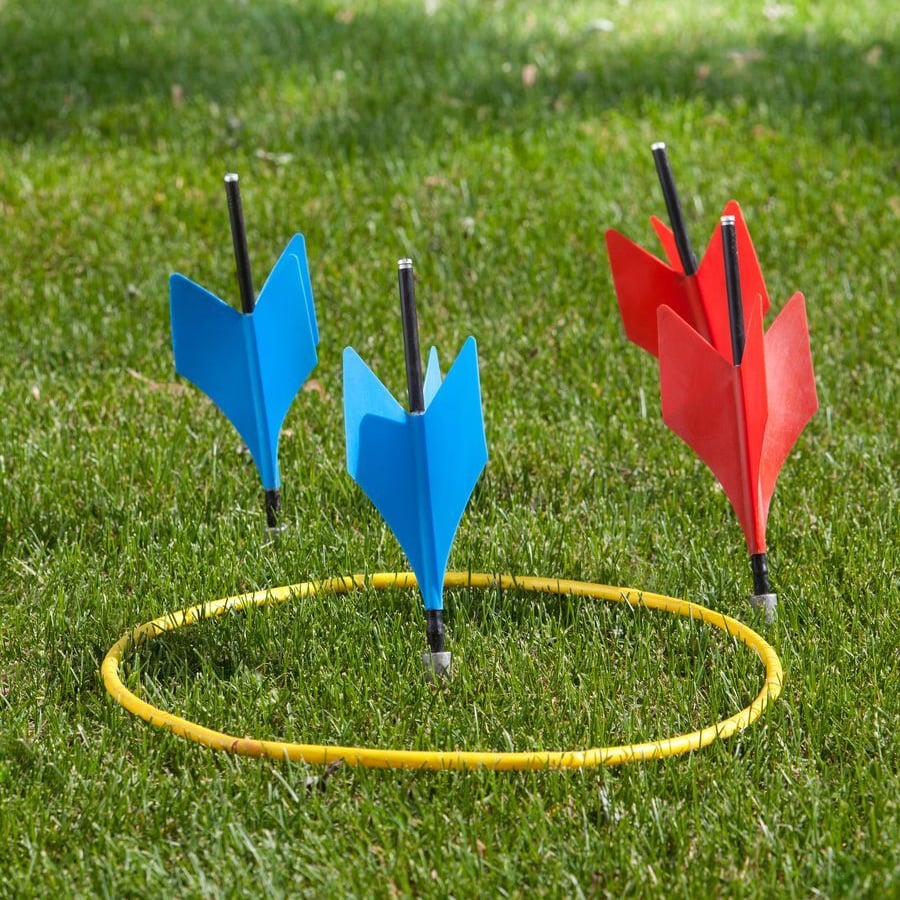 Four large plastic lawn darts, two blue and two red, are stuck in the grass near a yellow ring. The darts are standing upright, with two inside the ring and two outside, on a green lawn.